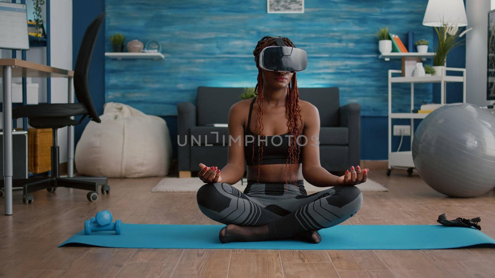 Fit black woman wearing virtual reality headset while sitting on yoga map in lotus position during meditation workout in living room. Athetic adult enjoying healthy lifestyle practicing sport