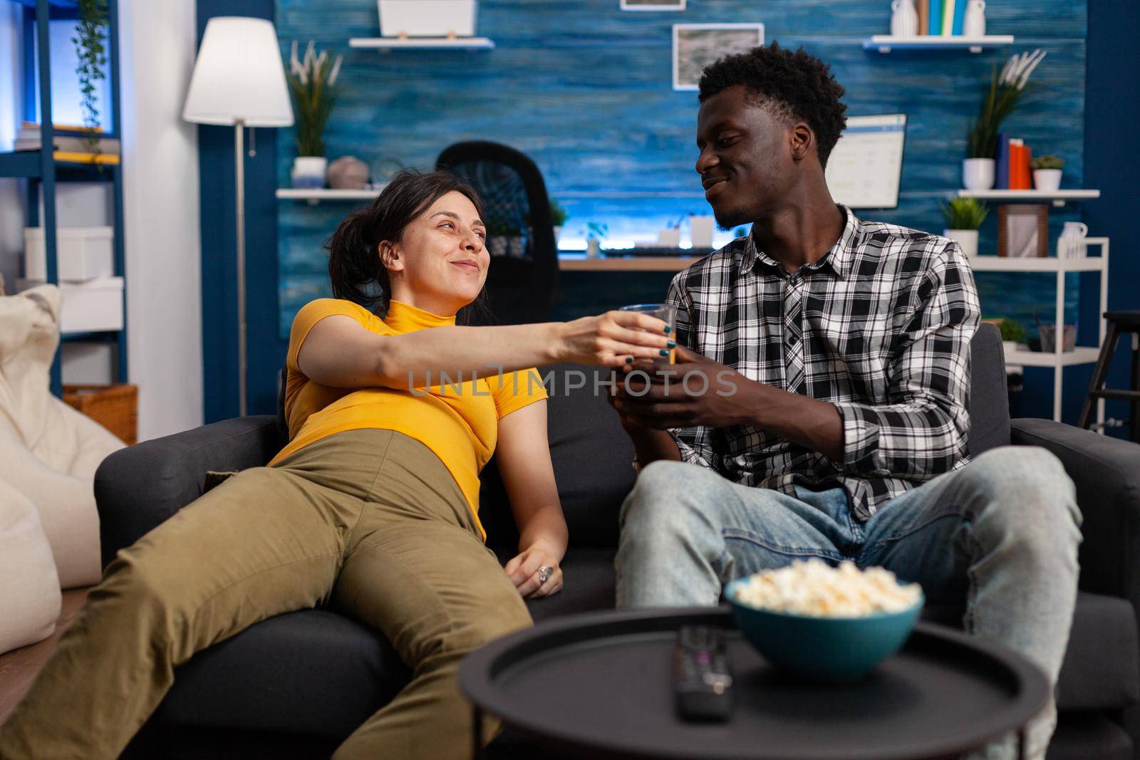 Interracial people with pregnancy sitting at home. African american father of child bringing glass of water for pregnant caucasian woman laying on couch. Mixed race couple expecting baby