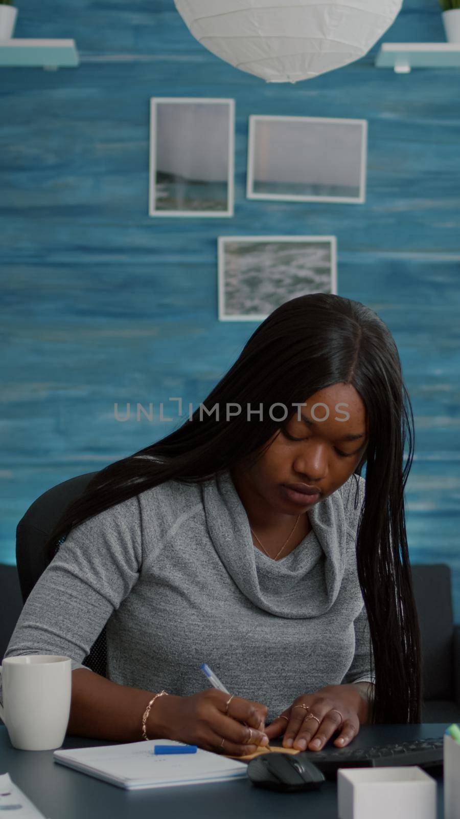 Student writing business ideas on sticky notes putting on computer working at school homework, using elearning platform during university online course. Black woman sitting at desk in living room