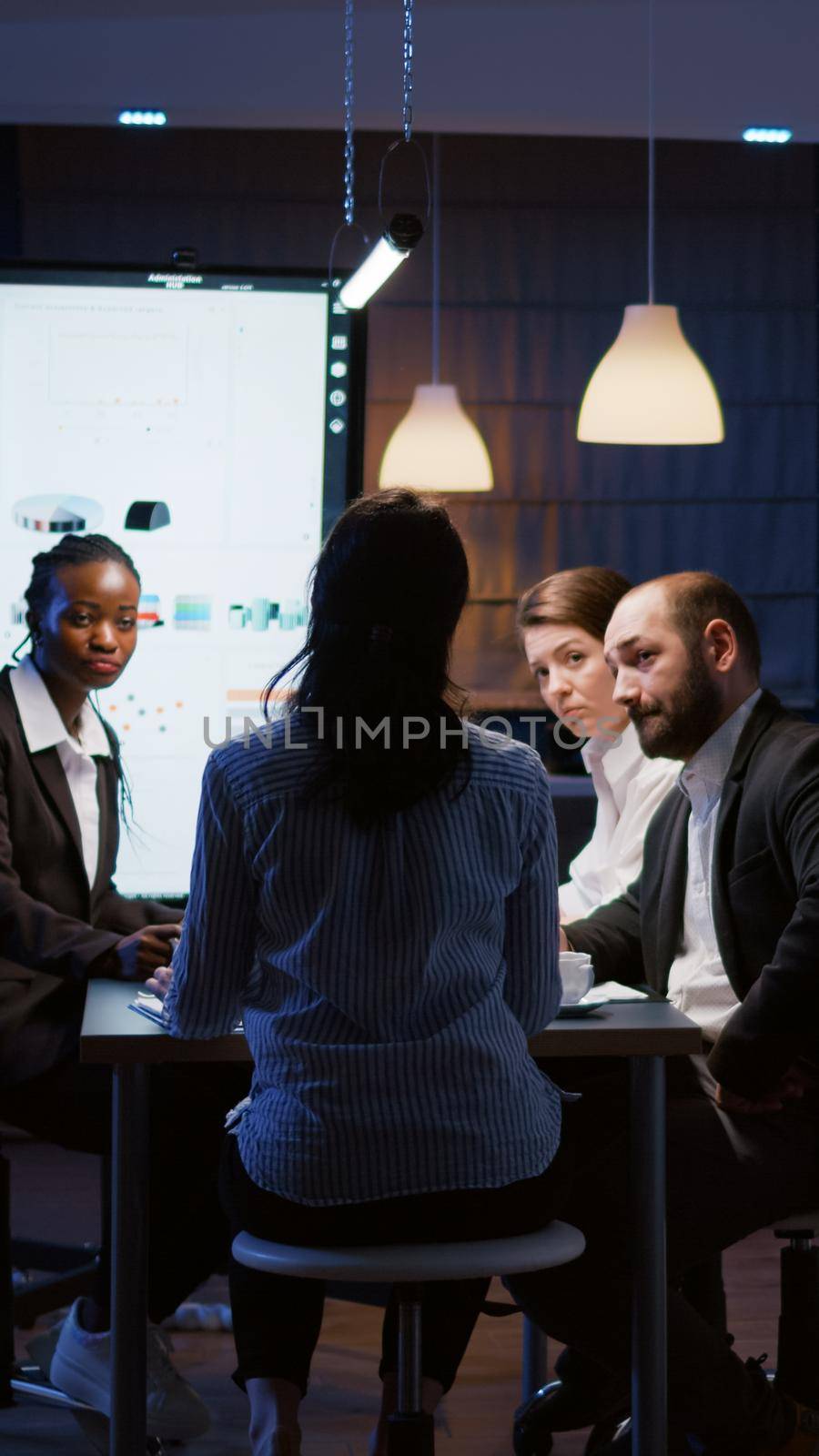 Selective focus of workaholic businesswoman back discussing financial graphs analyzing company paperwork overworking in office meeting room late at night. Multi-ethnic coworkes brainstorming ideas