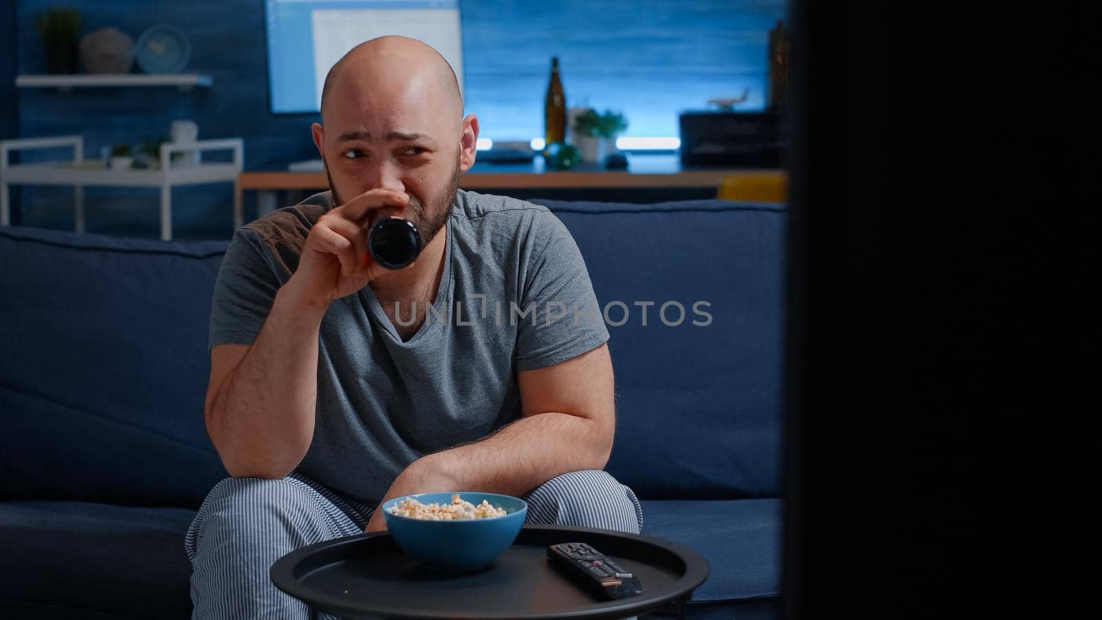 Man sitting on cozy couch in living room alone, eating popcorn, drinking beer choose comedy film for funny pastime, laughing at entertaining programs reality show series on tv for leisure