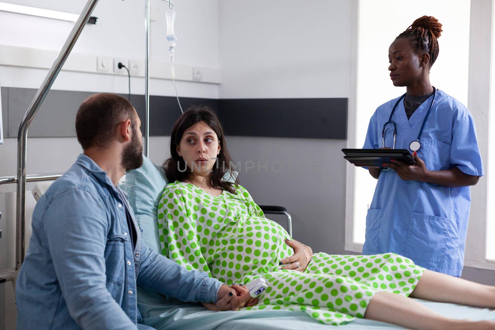 Young couple expecting child in hospital ward while talking to african american nurse about childbirth. Medical assistant consulting pregnant woman laying in bed and man holding hand