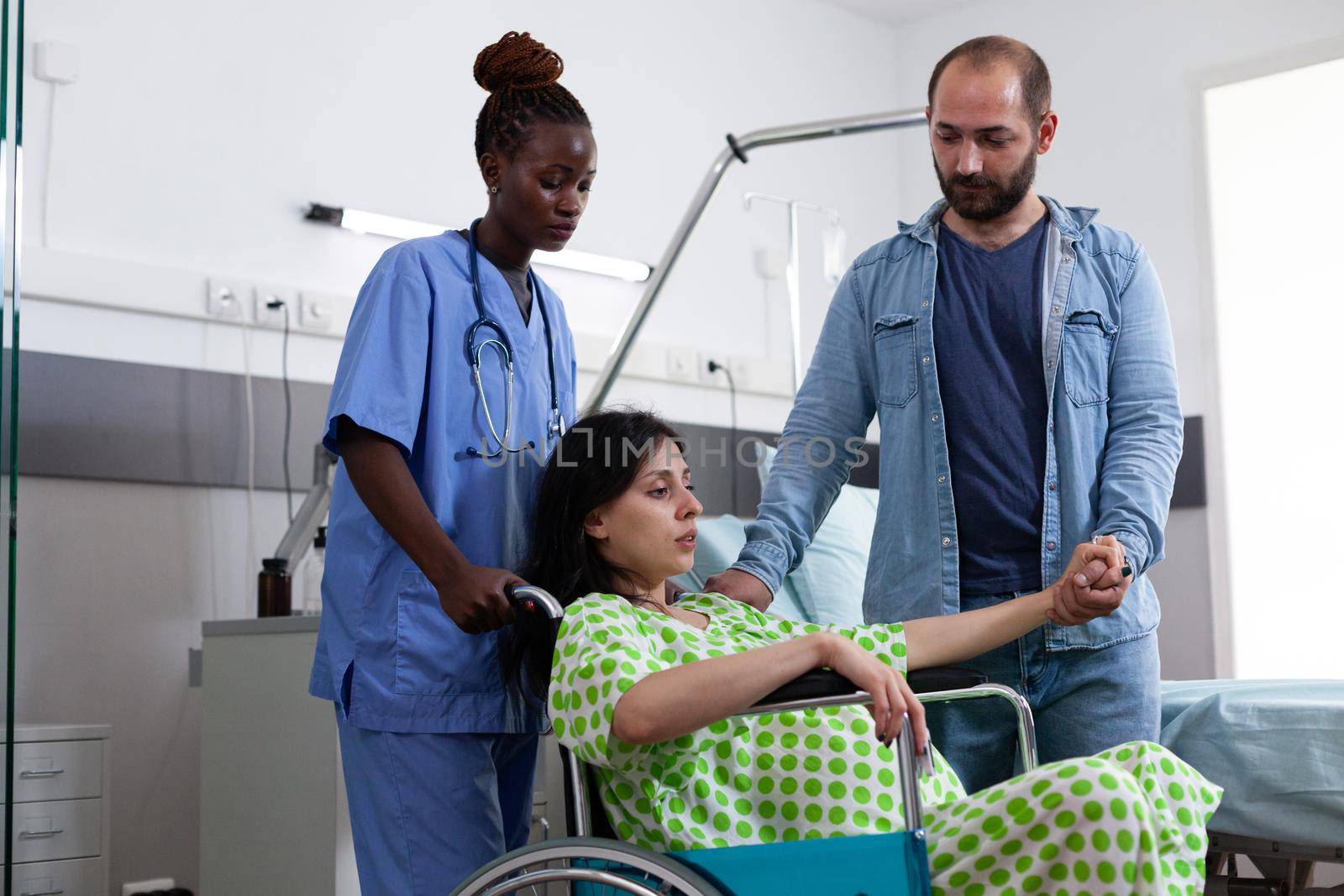 Black nurse transporting pregnant woman in wheelchair to maternity room for child delivery. Person with baby bump preparing for childbirth while holding hand of husband in hospital ward