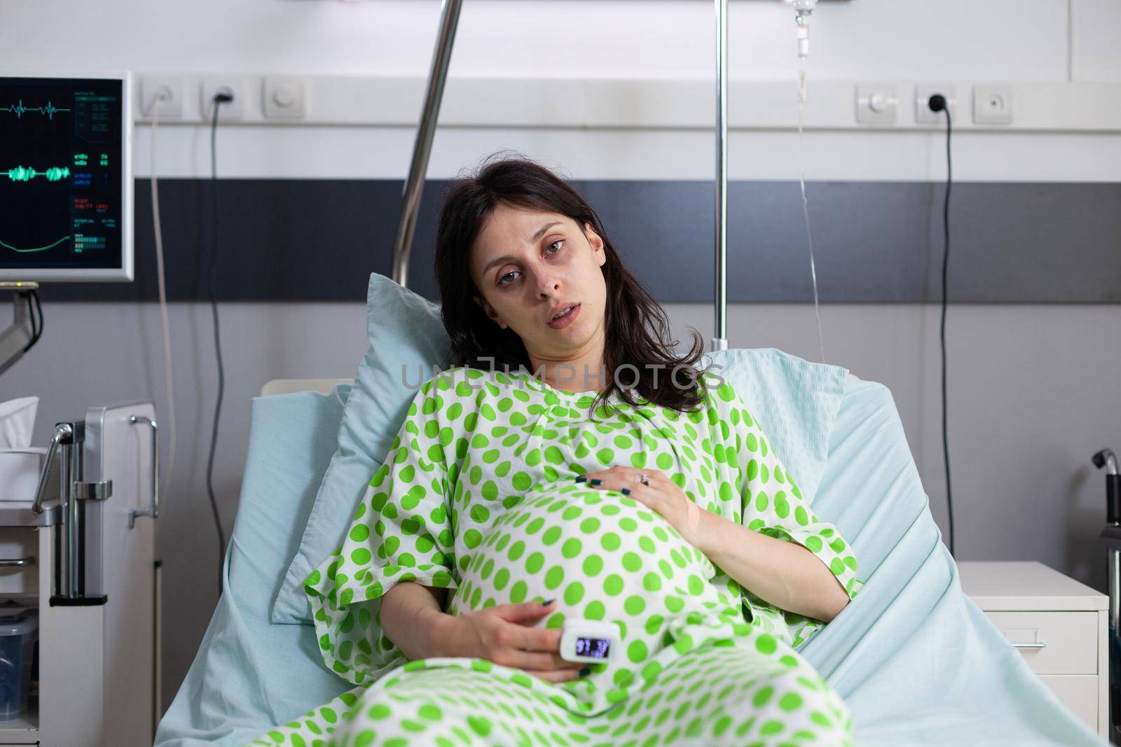 Portrait of tired pregnant person sitting in hospital ward bed at medical facility. Woman expecting child, looking at camera and holding hands on belly. Young adult with pregnancy