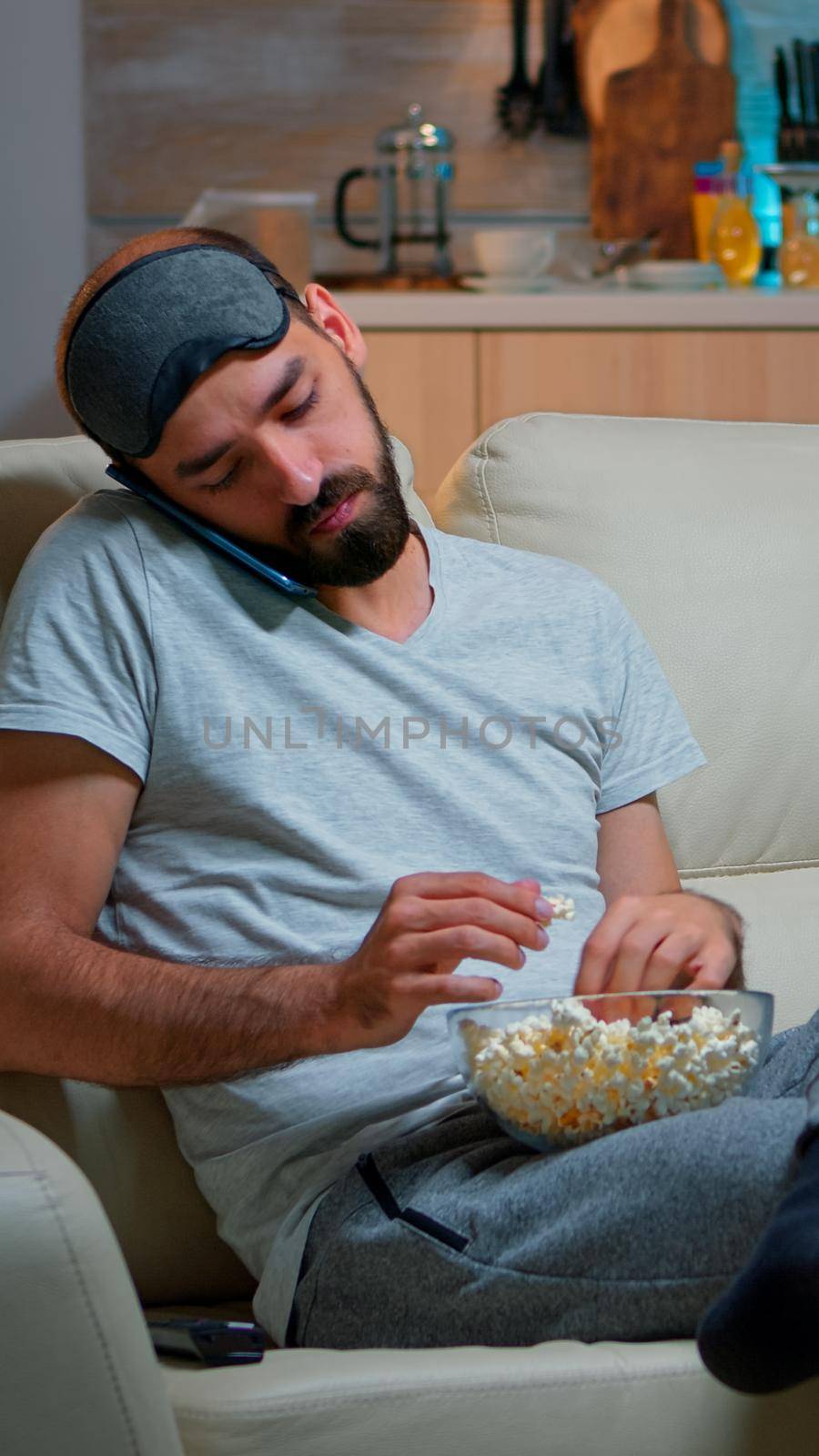 Relaxed man talking about internet connection on smartphone. Caucasian male sitting on couch watching entertainment movies while eating popcorn late at night in kitchen