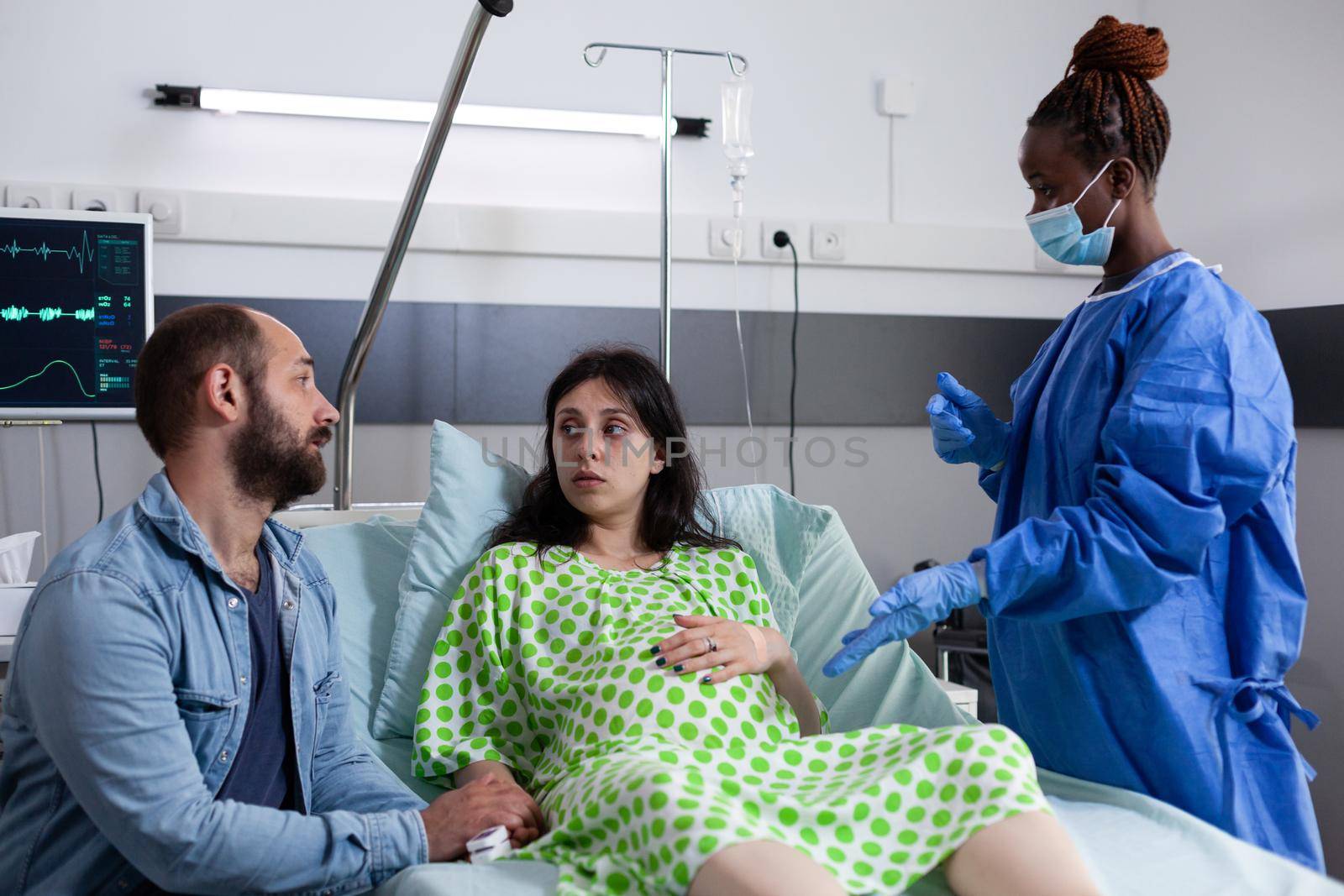 Young parents expecting child at medical clinic, discussing childbirth with african american nurse while pregnant woman sitting in hospital ward bed. Caucasian couple with pregnancy