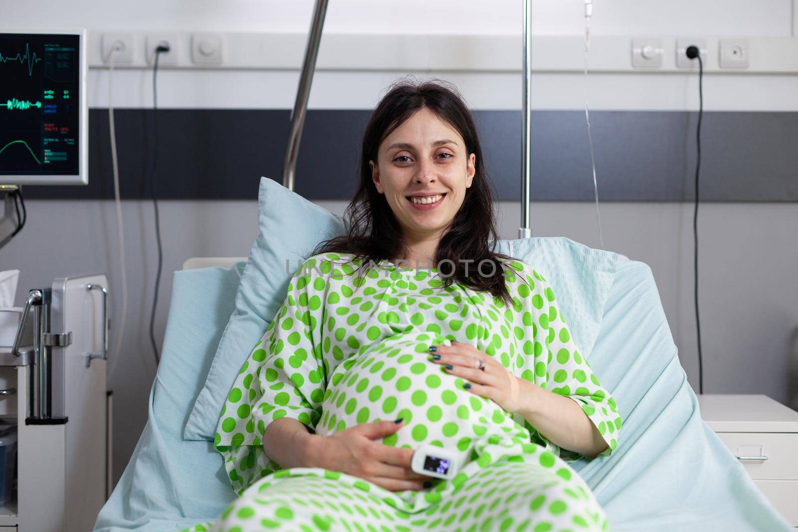 Portrait of pregnant woman sitting in hospital ward bed, looking at camera. Young person smiling, waiting for child delivery and parenthood at medical clinic. Adult with baby bump