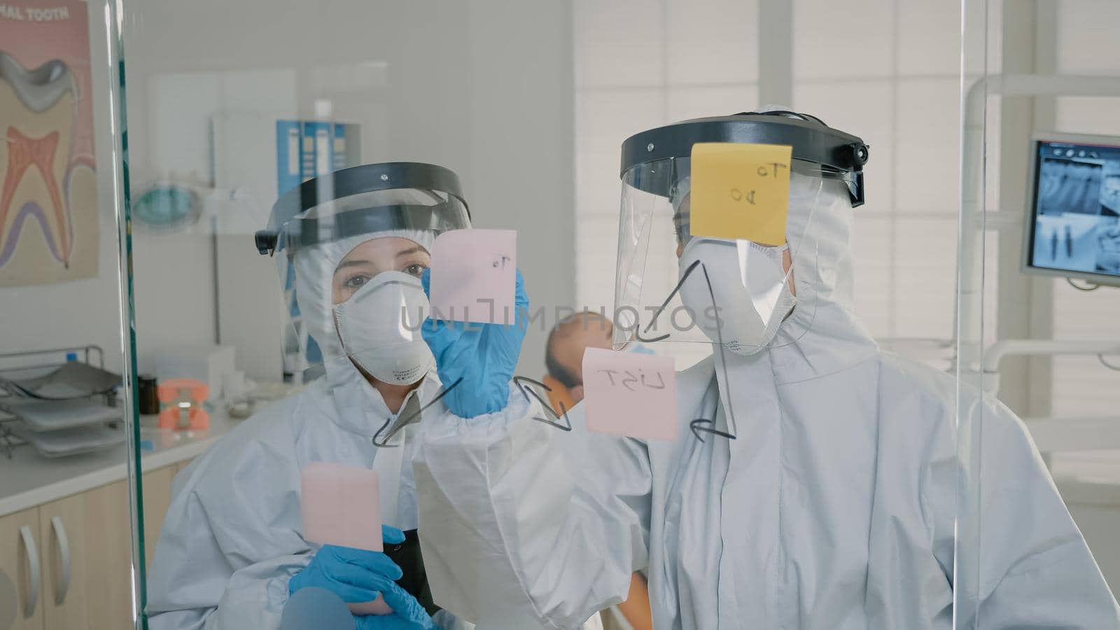 Orthodontists in ppe suits making diagram with sticky notes on glass at dentistry office. Dentist and dental assistant using tools for oral care operation on patient sitting in cabinet
