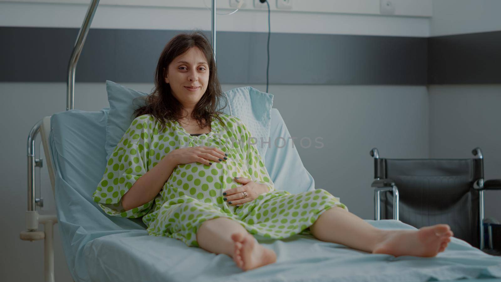 Portrait of pregnant caucasian woman sitting in hospital ward bed at healthcare clinic. Impatient person with pregnancy baby bump preparing for child delivery and medical assistance