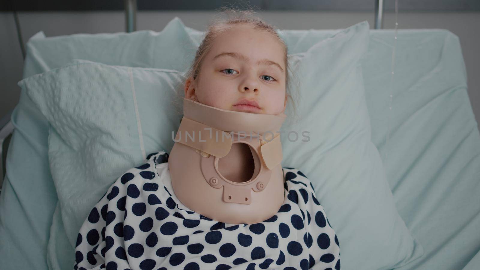 Portrait of sick little patient resting in bed looking at camera while having neck cervical collar recovering after painful surgery in hospital ward. Child wearing oxygen nasal tube during examination