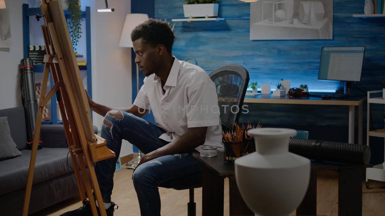 African american adult making fine art sitting in artwork studio with artistic tools. Black young man designing drawing of vase with pencil and white canvas on easel. Person with creativity