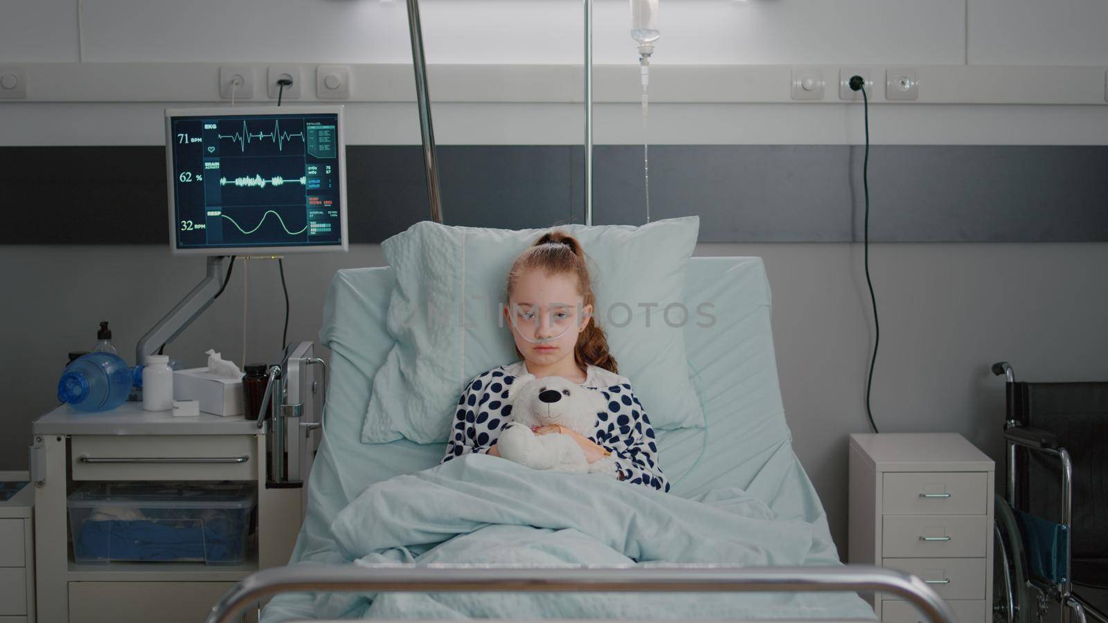 Portrait of hospitalized sick girl child patient holding teddy bear resting in bed during medical consultation in hospital ward. Alone kid wearing oxygen nasal tube waiting for disease examination