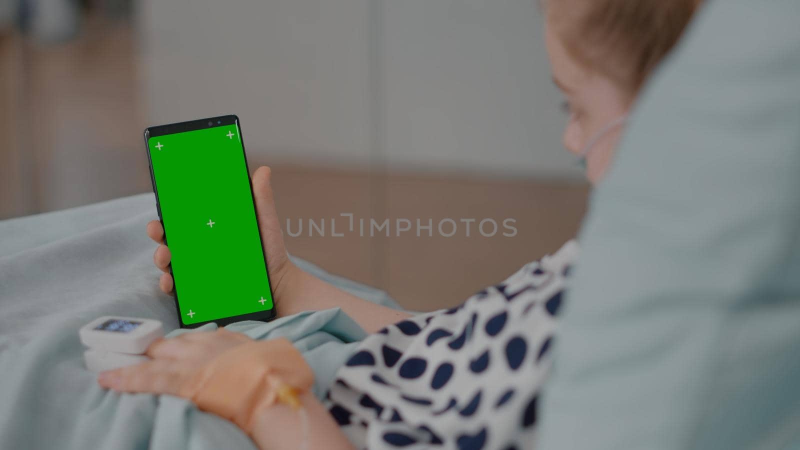 Sick girl resting in bed with medical oximeter on finger looking at green screen mock up chroma key phone with isolated display. Child suffering recovery surgery during examination in hospital ward