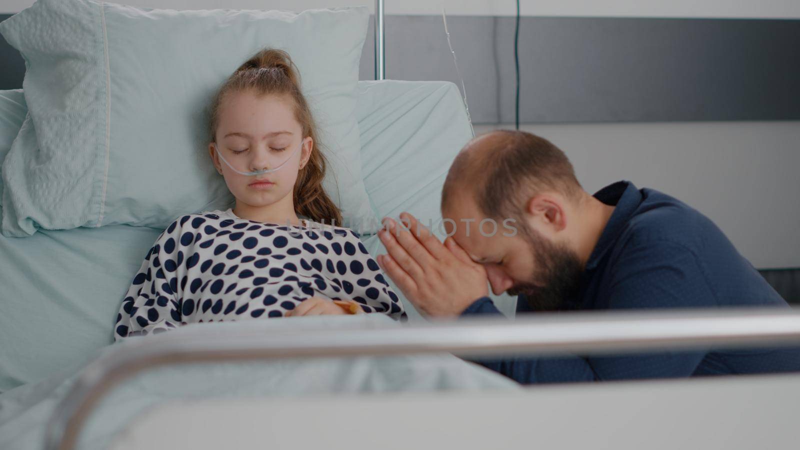 Worried father praying for sick girl daughter health recovery after suffering medical surgery in hospital ward. Little child with oxygen nasal tube sleeping during sickness examination