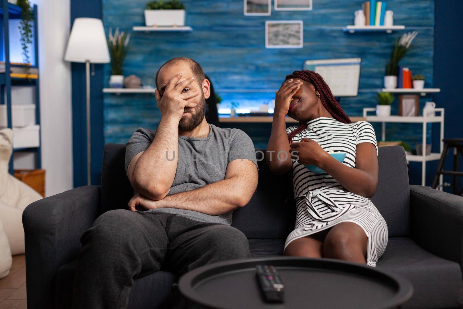 Interracial couple watching scary movie on television and covering eyes, being afraid. Multiracial partners feeling scared while looking at camera and horror movie on TV sitting at home