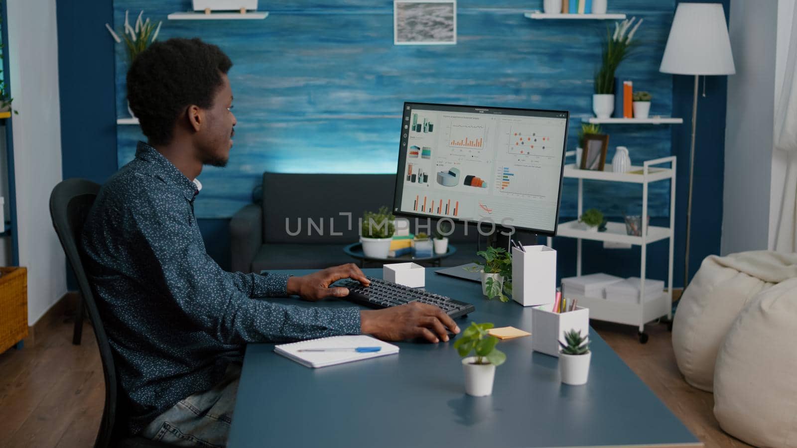 African american guy working from home, taking notes on notepad while remote working on computer from living room. Computer user using business internet online web communication job