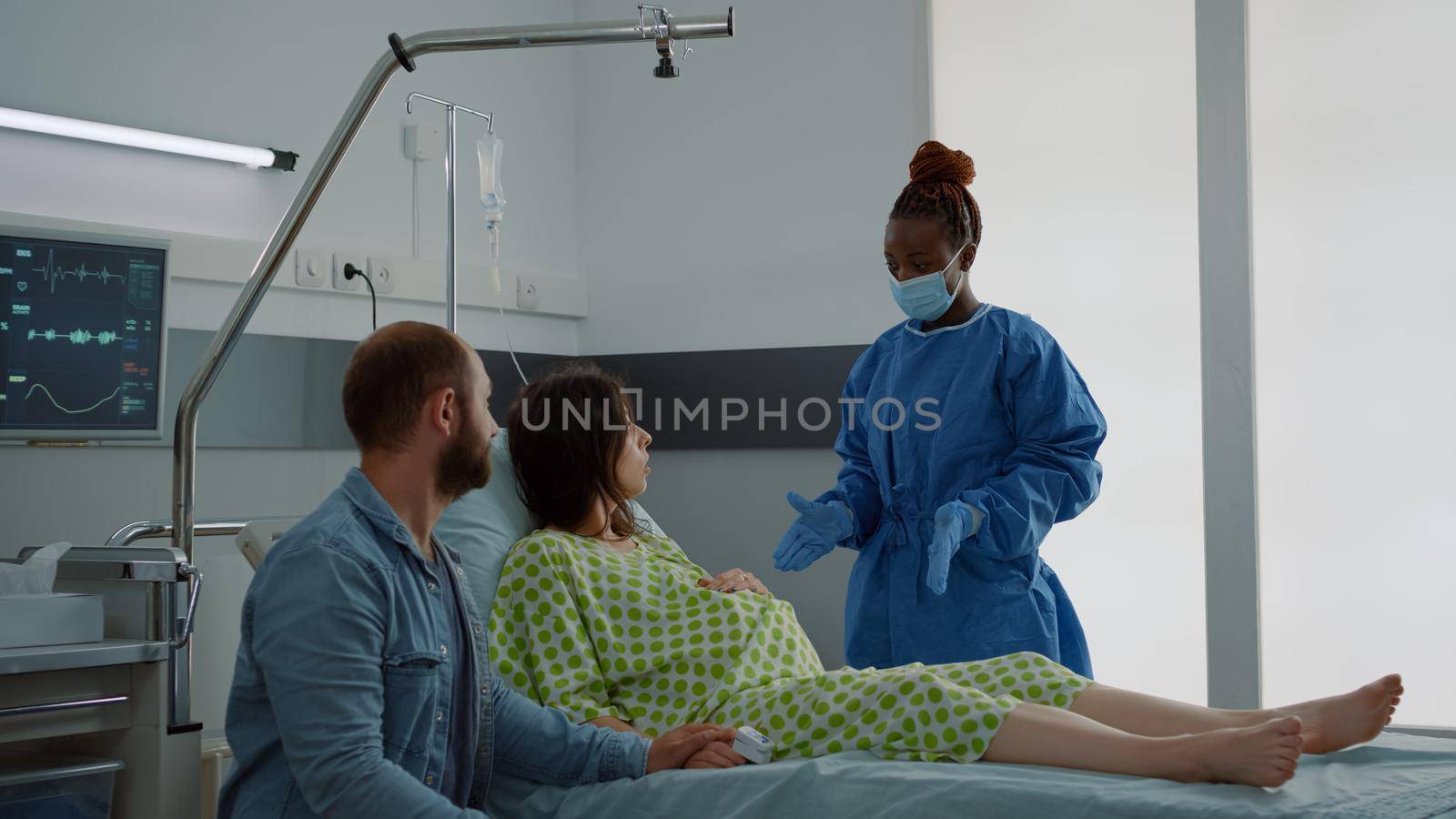 Wife expecting child sitting in hospital ward with husband waiting for delivery preparations at maternity clinic. Young caucasian couple talking to african american nurse about childbirth