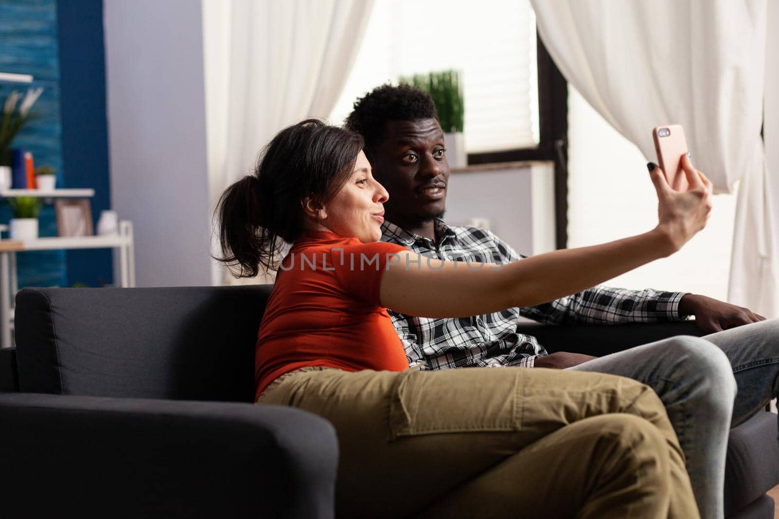 Cheerful interracial couple taking pictures with smartphone at home. Multi ethnic people making memories and selfies with modern device in hand and technology. Mixed race happy partners