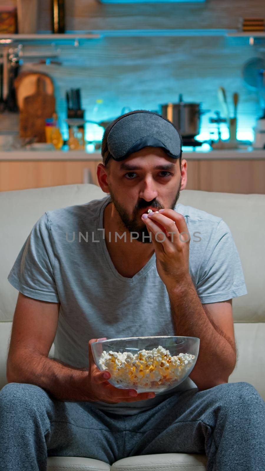 Concentrated man with sleep eye mask sitting in front of television watching lifestyle show. Caucasian male relaxing on couch holding popcorn bowl late at night in kitchen