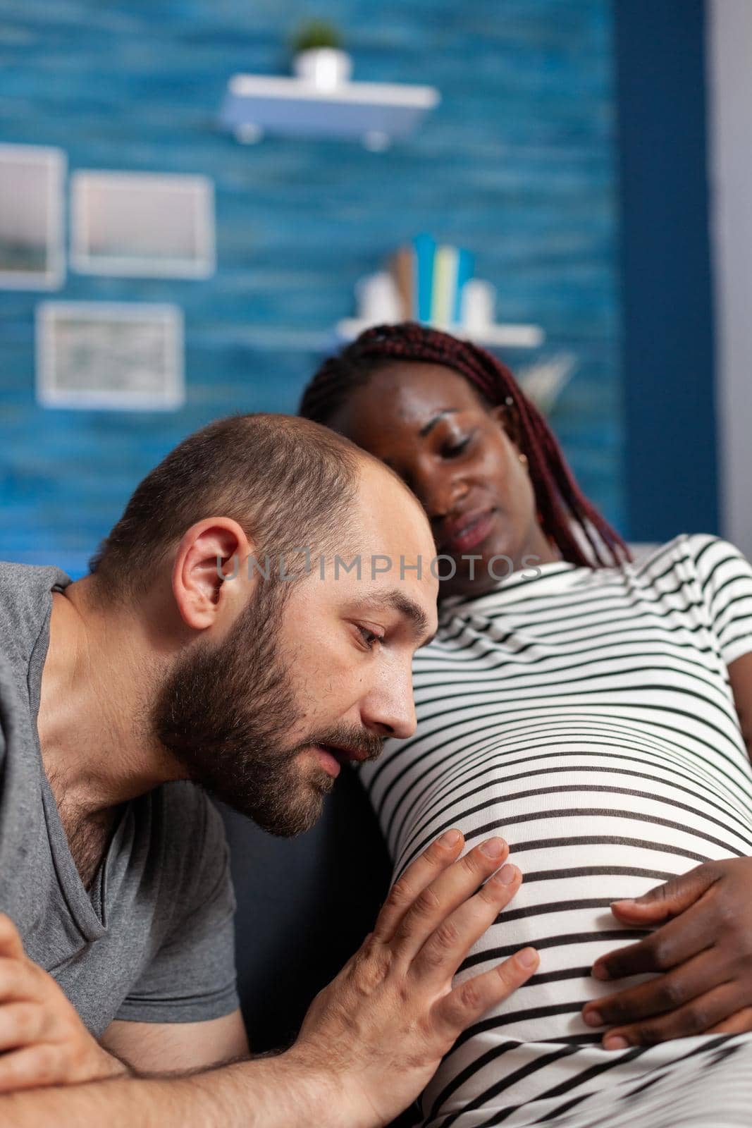 Close up of caucasian father of baby looking at belly and holding hand while pregnant black woman smiling. Interracial couple with pregnancy expecting child and relaxing at home