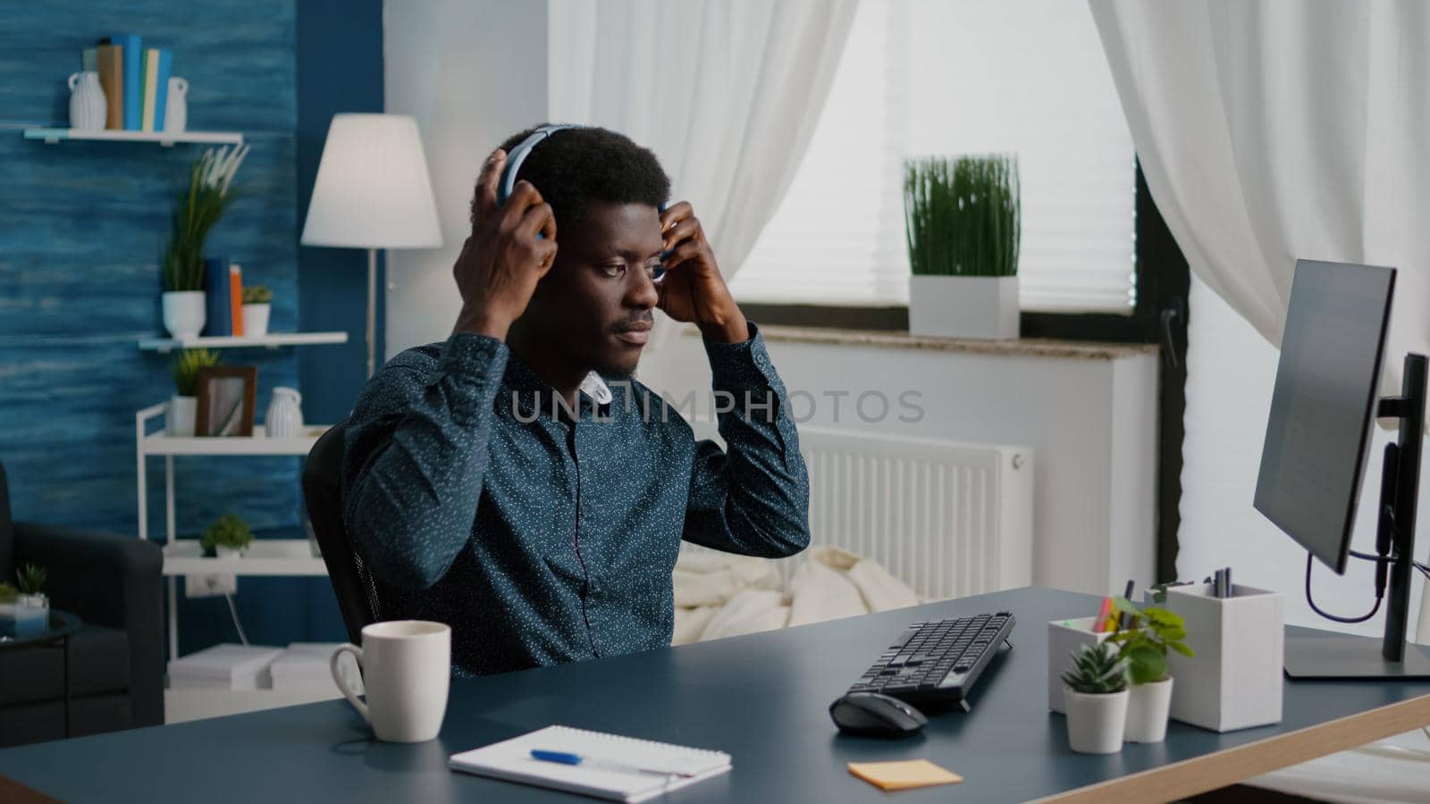 Black freelancer of african ethnicity putting headphones on while working from home, remote worker computer user in home office typing on PC listening music. Concentrated guy enjoying music