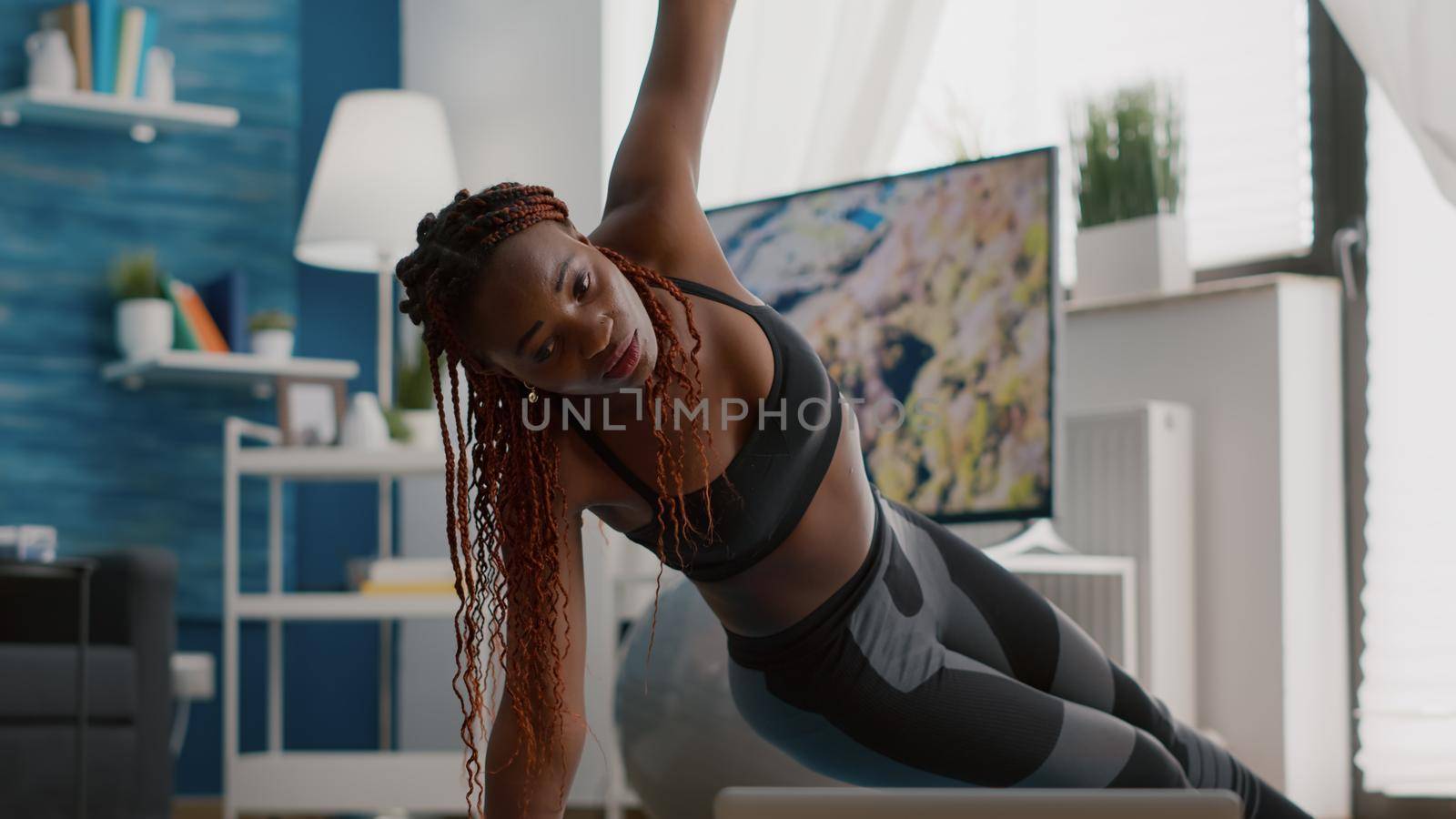 Athlete black woman training on yoga map in living room standing in side plank watching fitness video using laptop computer. Adult in sportwear streching muscle during strech gymnastics