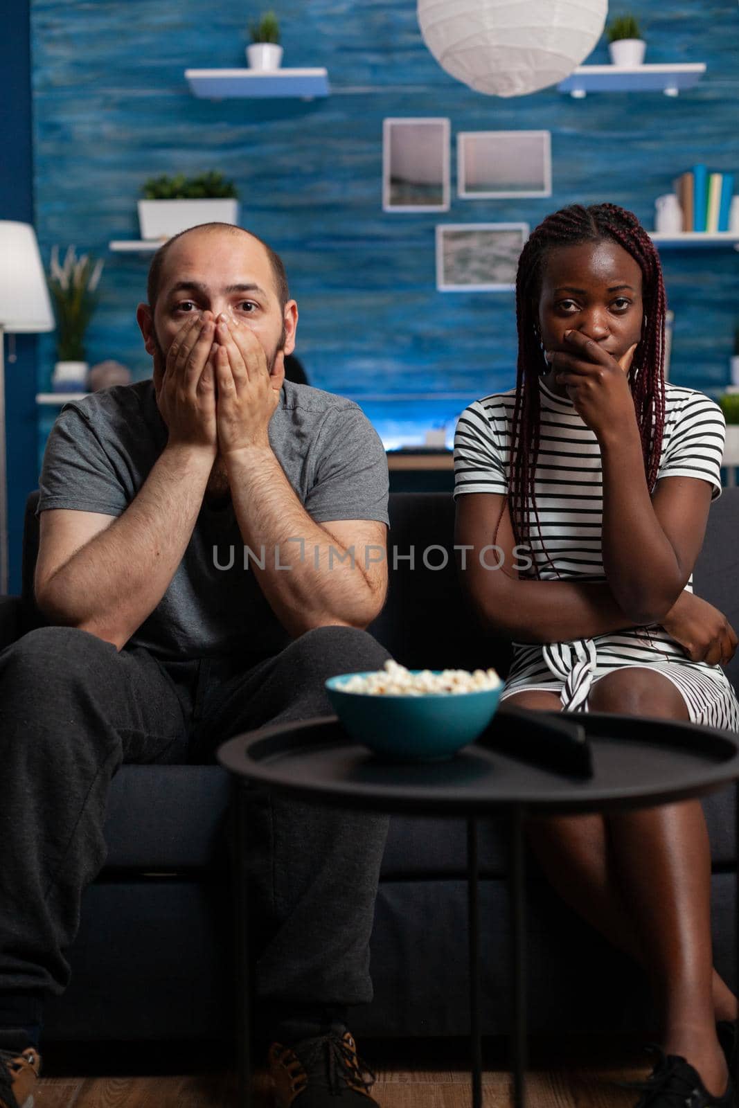 POV of interracial couple being shocked watching drama movie on television in living room. Mixed race partners with hands over mouth looking at camera and TV. Multi ethnic lovers