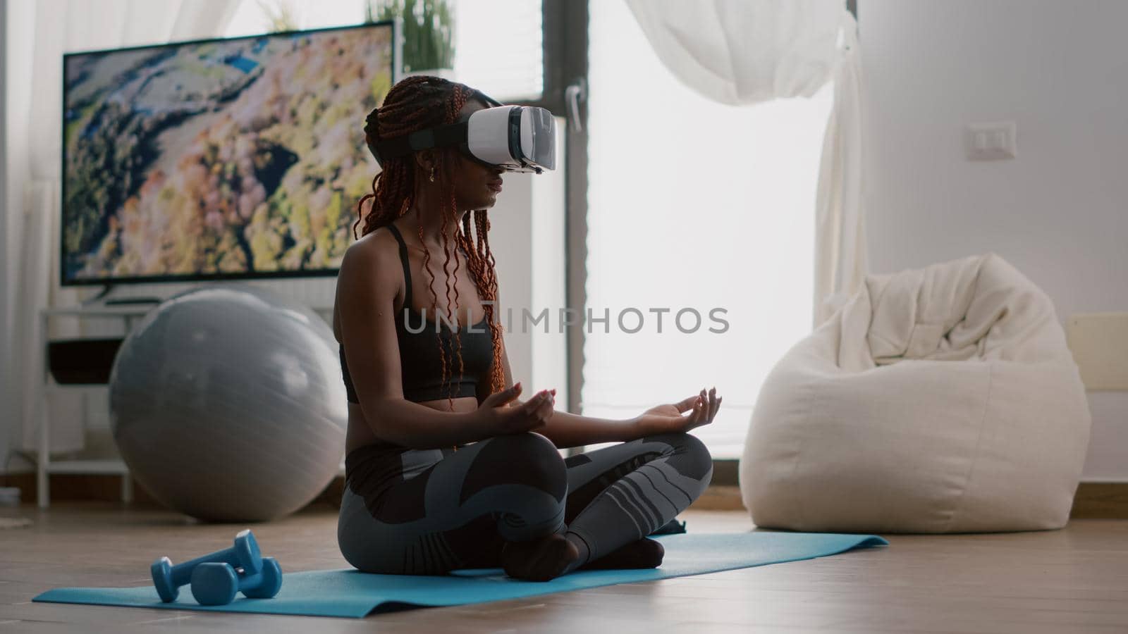 Black trainer woman wearing virtual reality headset while sitting on yoga map in lotus position during meditation workout class in living room. Athetic adult enjoying healthy lifestyle training body muscle