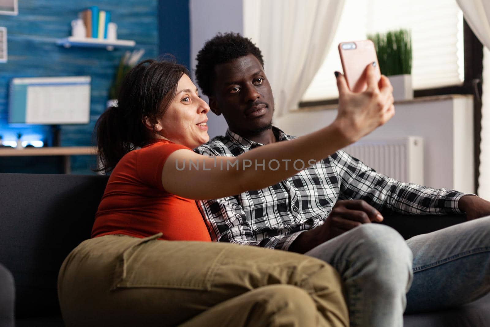 Cheerful interracial couple taking pictures with smartphone at home. Multi ethnic adults creating memories and selfies with modern gadget in hand and technology. Mixed race happy partners