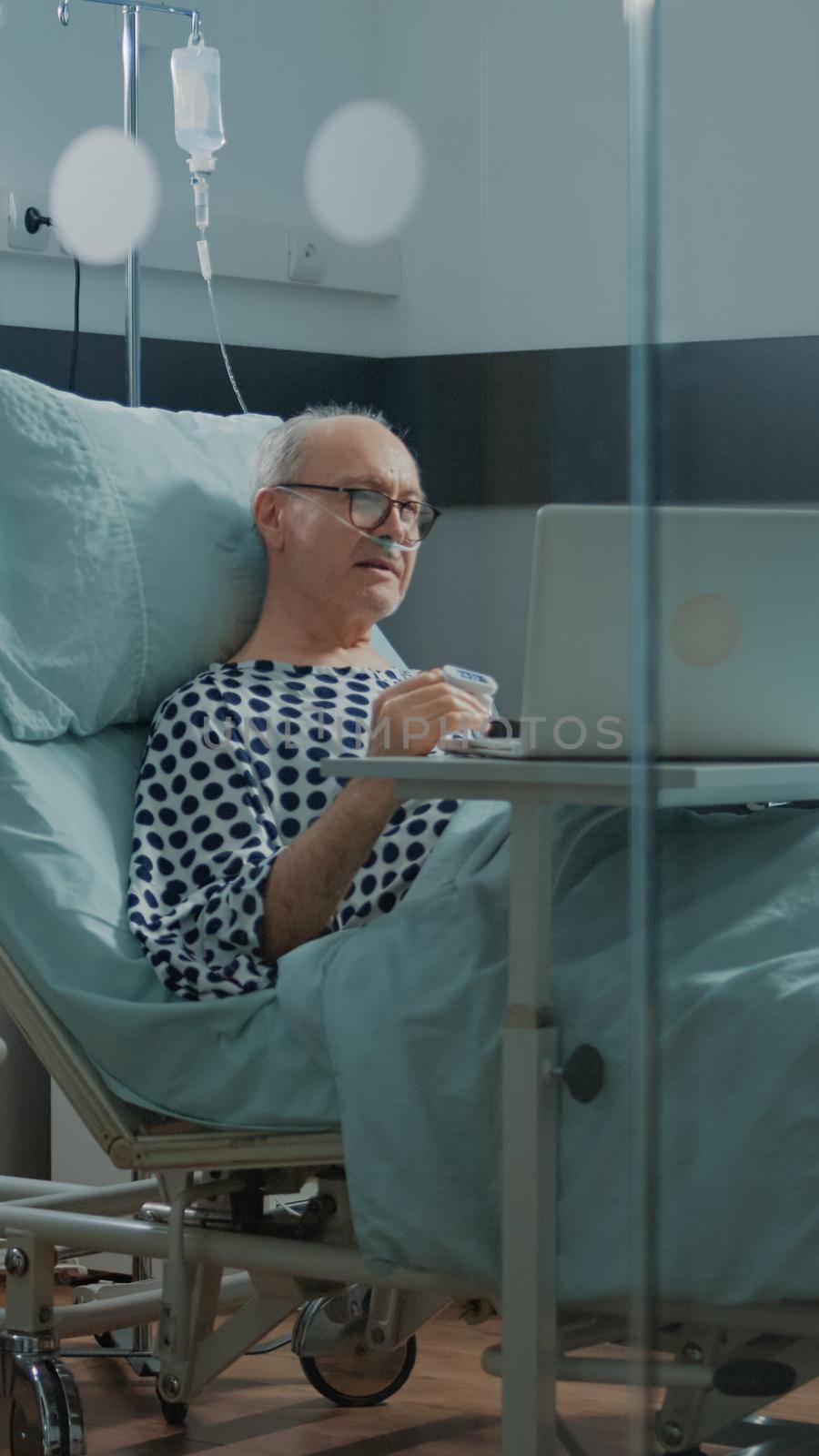 Elder sick patient using laptop in hospital ward bed to talk to relatives about health problems. Old man on videocall waiting for healing treatment to cure pain from surgery and disease