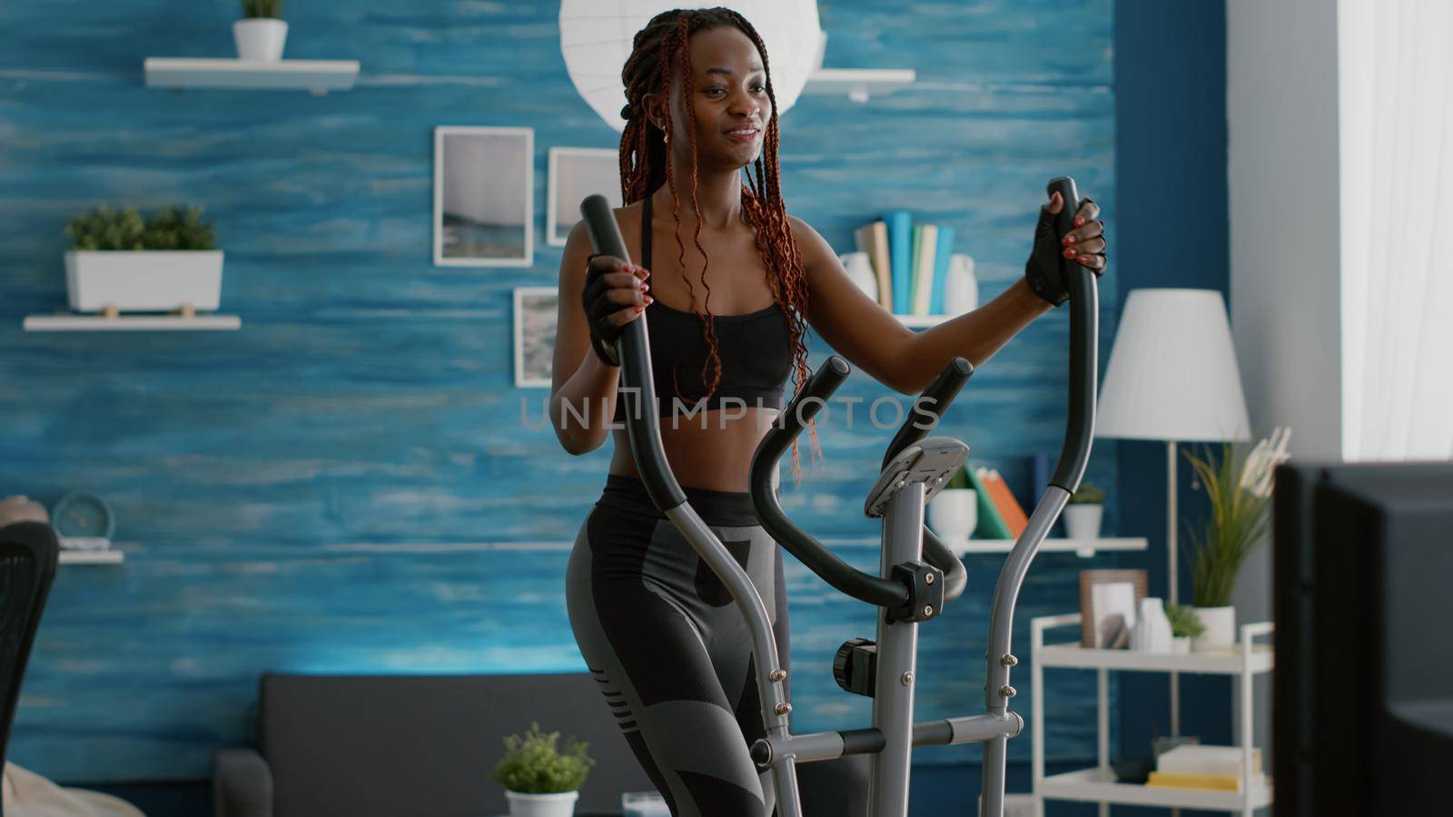 Black athletic woman doing cardio training on elliptical bike in living room during cardio morning routine in living room. Trainer in sportwear warming up bodyweight enjoying healthy lifestyle
