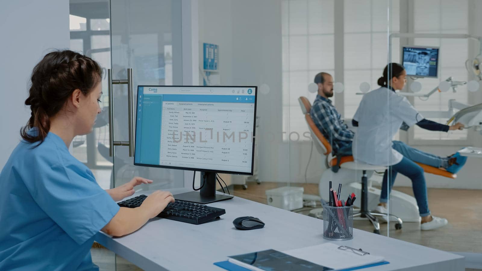 Stomatology nurse sitting at desk working on computer for appointments at dental clinic. Assistant using monitor screen while dentist doing professional examination in background