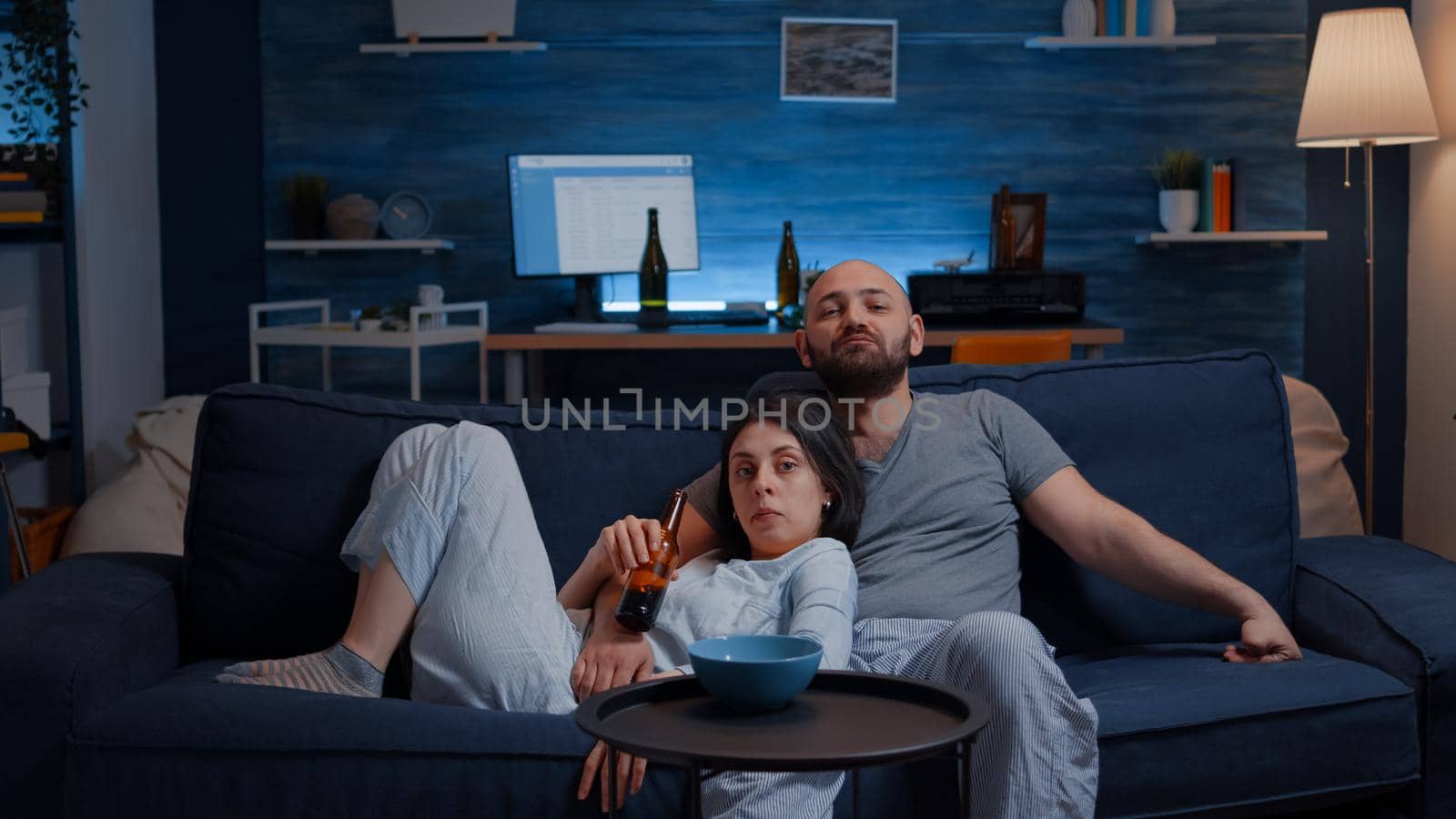Relax couple in pajamas sitting on sofa eating popcorn watching TV, enjoying free time together looking at entertainment show sitting on confortable couch. Husband and wife relaxing with comedy movie