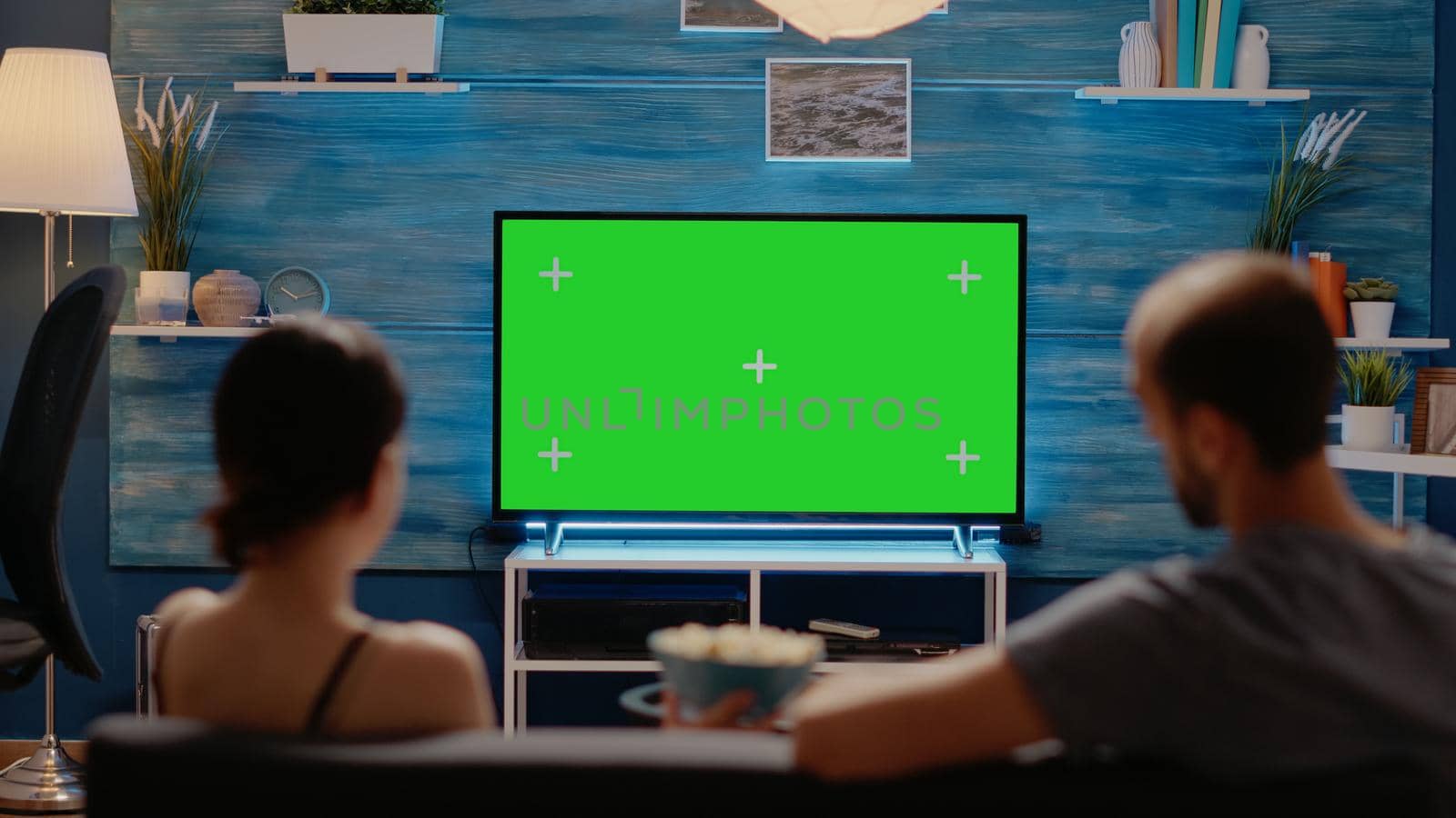 Young man and woman watching green screen on tv display sitting on couch at home. Caucasian people using chroma key and modern equipment on copy space for mockup and isolated template