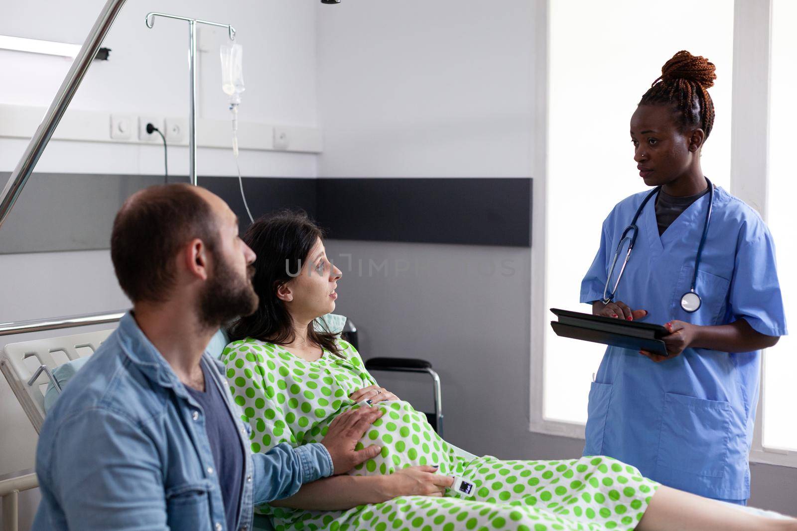 Caucasian couple expecting child in hospital ward while talking to african american nurse at maternity. Parents with pregnancy receiving medical assistance preparing for child delivery