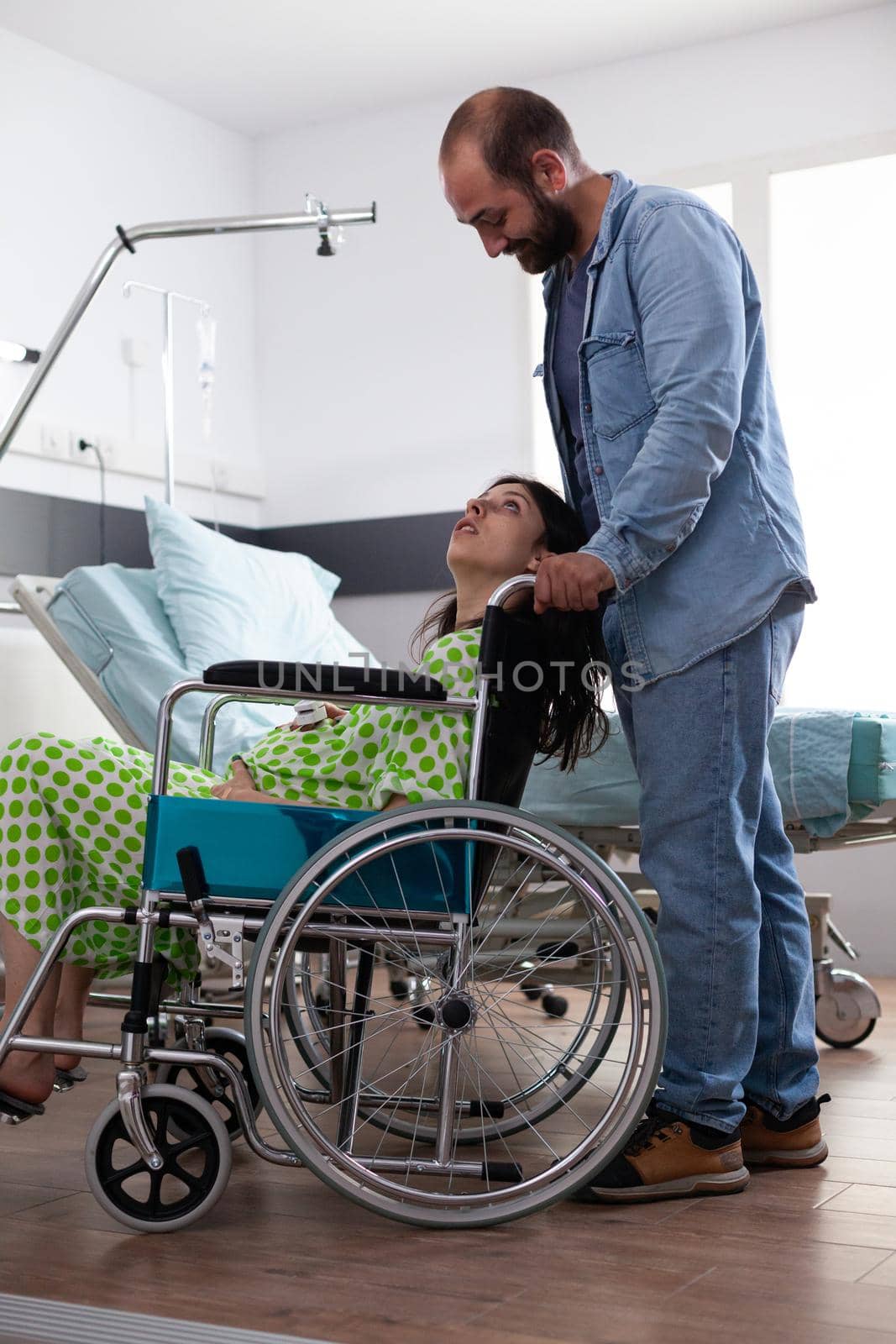 Caucasian pregnant woman sitting in wheelchair while father of baby assisting wife preparing for child delivery in hospital ward at medical facility. Young couple getting ready for childbirth