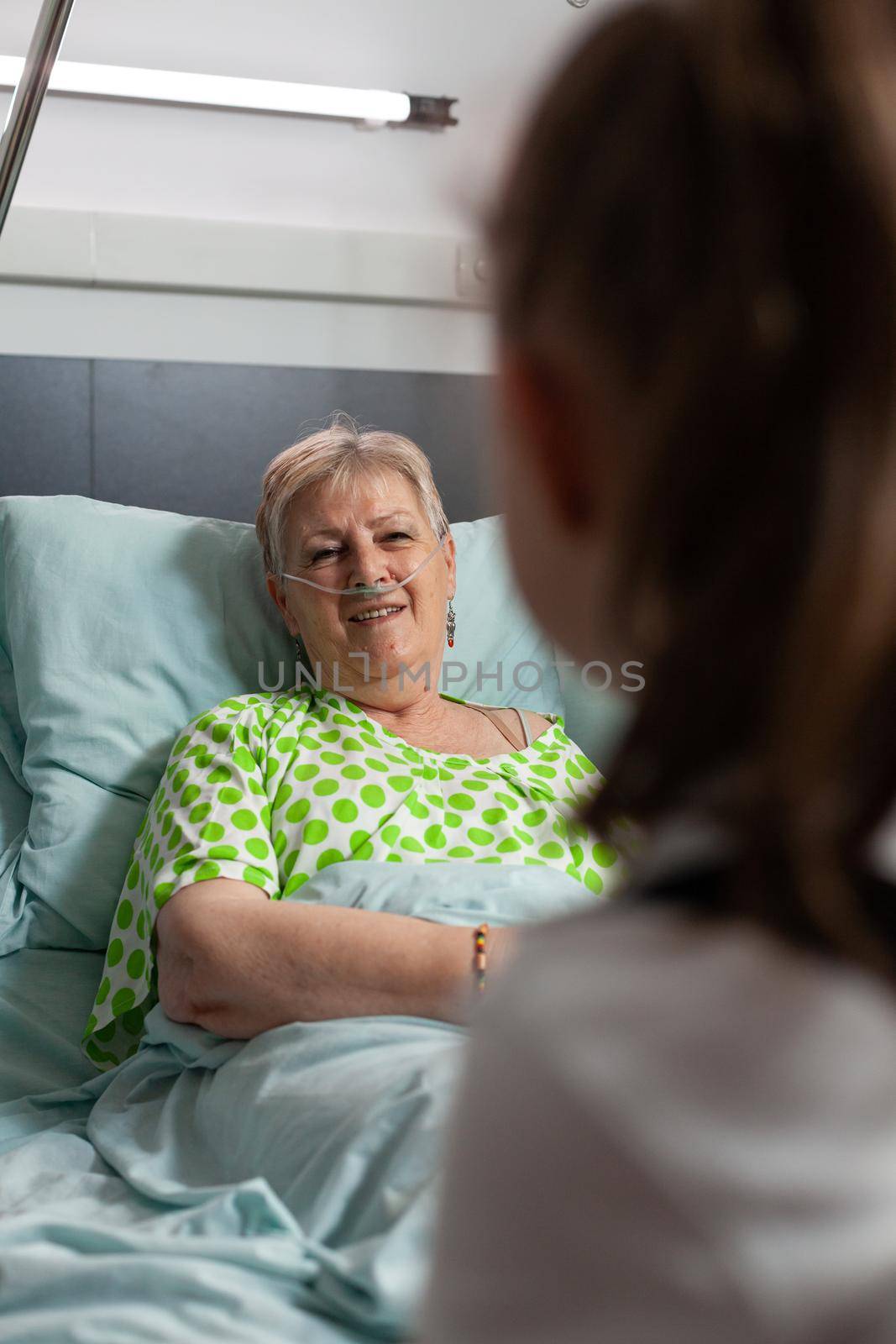Sick retired grandmother talking with grandchild while resting in bed after medical surgery in hospital ward. Family visiting grandparent supporting during illness examination. Healthcare therapy