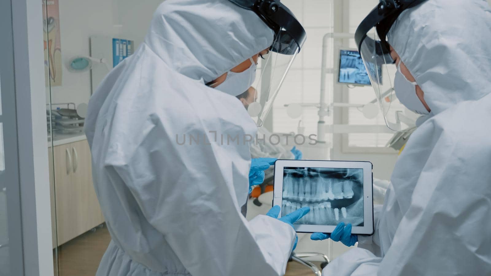 Professional dentists using technology for oral examination while wearing protection suits at dental clinic. Stomatology doctors discussing and looking at x ray scan on modern tablet