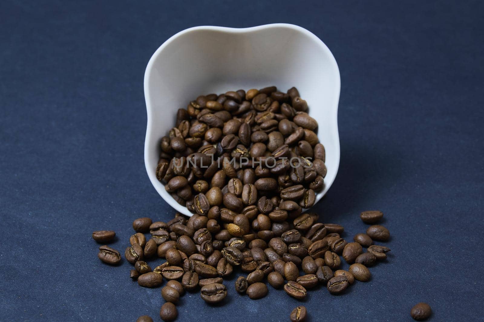 brown coffee beans roasted on a black background spilled out of an upturned white cup on the table