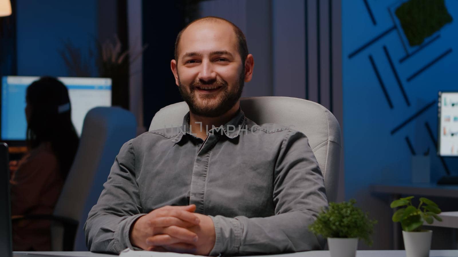 Portrait of smiling businessman sitting at desk table in business company office late at night looking into camera. Executive manager working at marketing statistics planning financial strategy