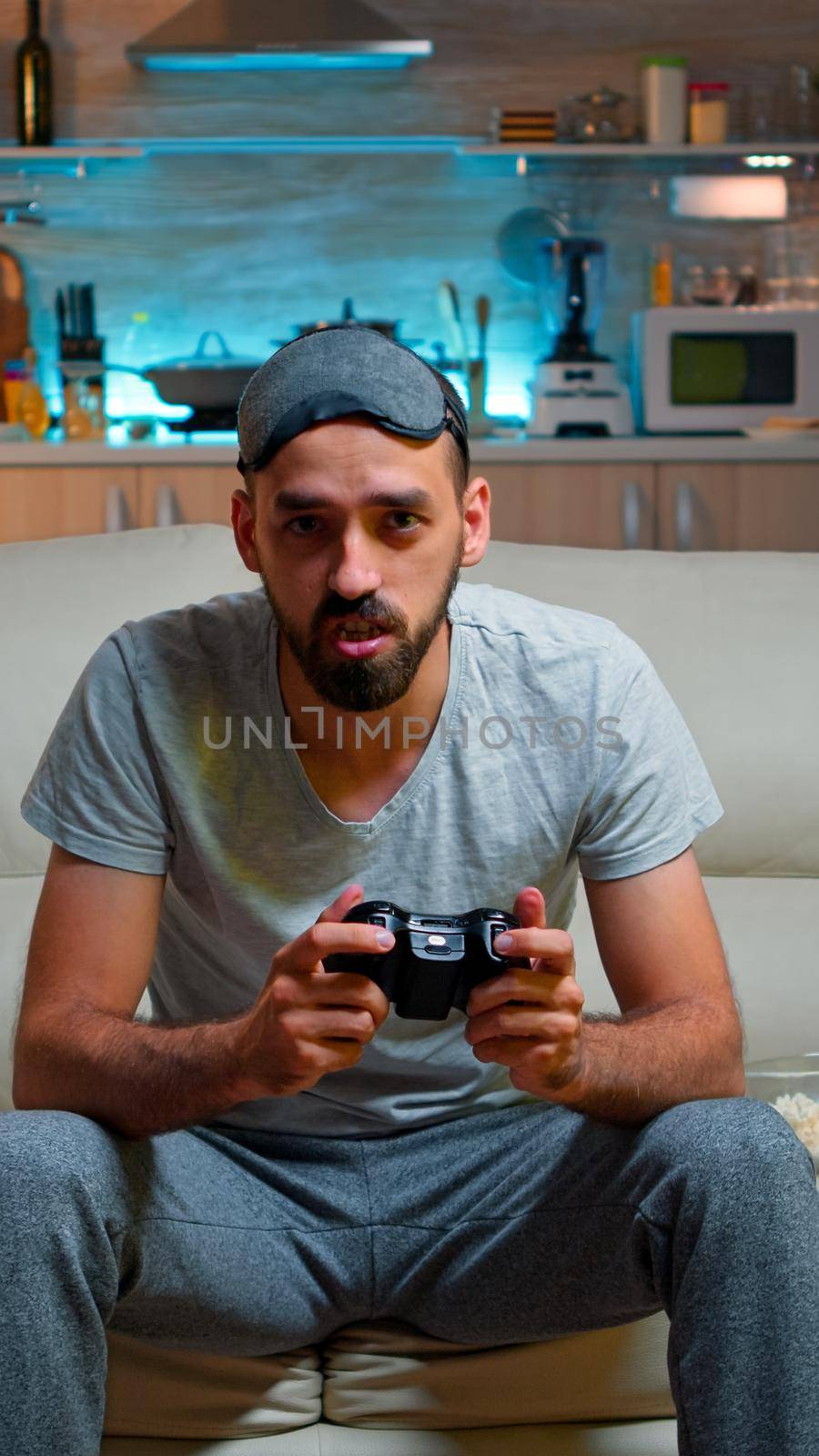 Focused man holding joystick while sitting in front of television winning online video games competition. Caucasian male with sleep mask lying on couch making winner gesture late at night in kitchen