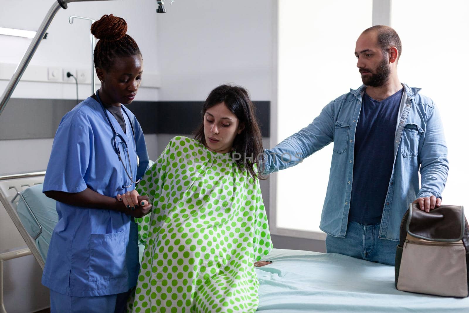 Black obstetrics nurse helping pregnant woman get out of bed in hospital ward at facility. Person with pregnancy belly receiving support from african american assistant and father of child