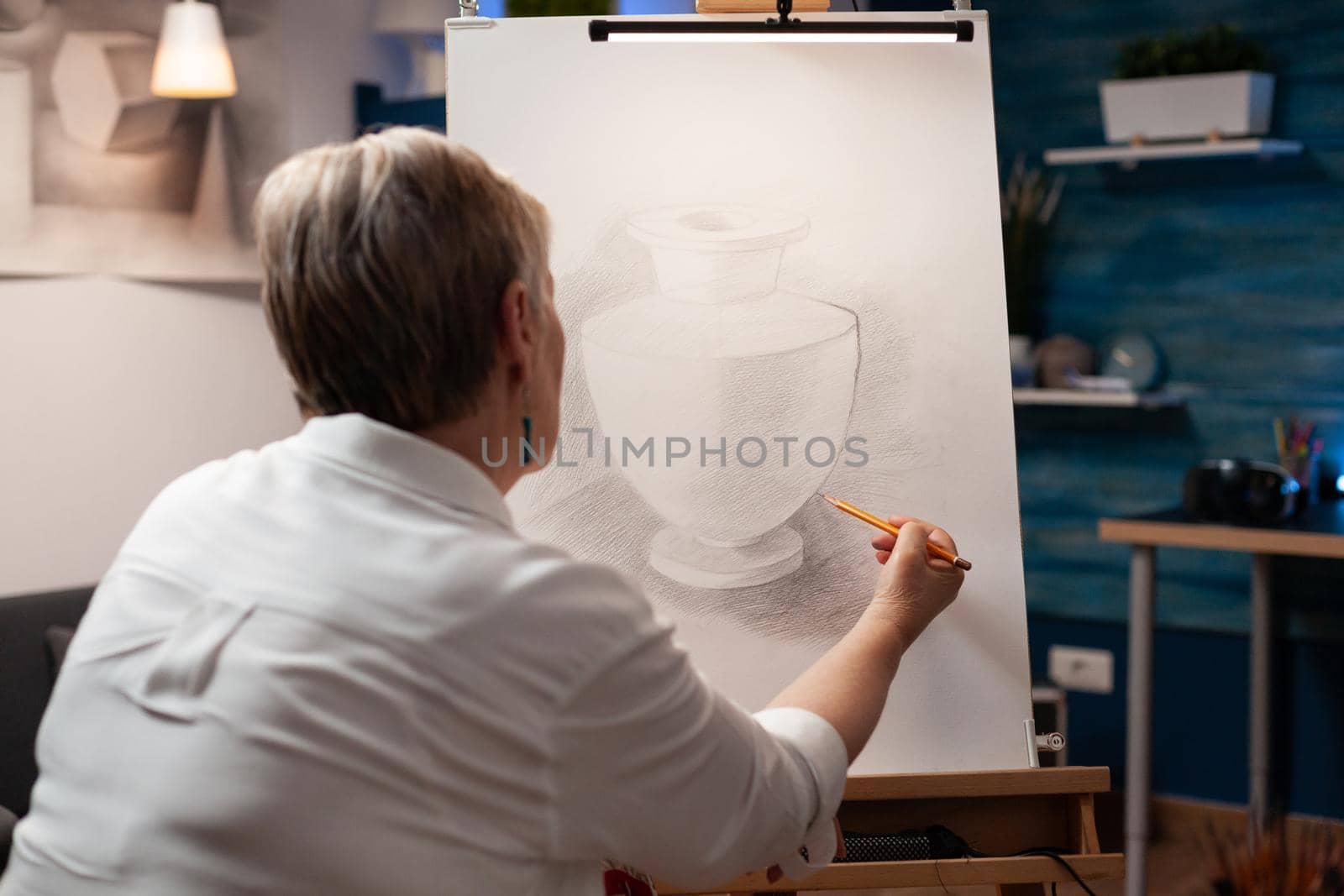 Close up of elderly woman drawing vase on canvas with pencil in workshop studio. Caucasian senior artist using object and artistic intruments for masterpiece artwork creation.