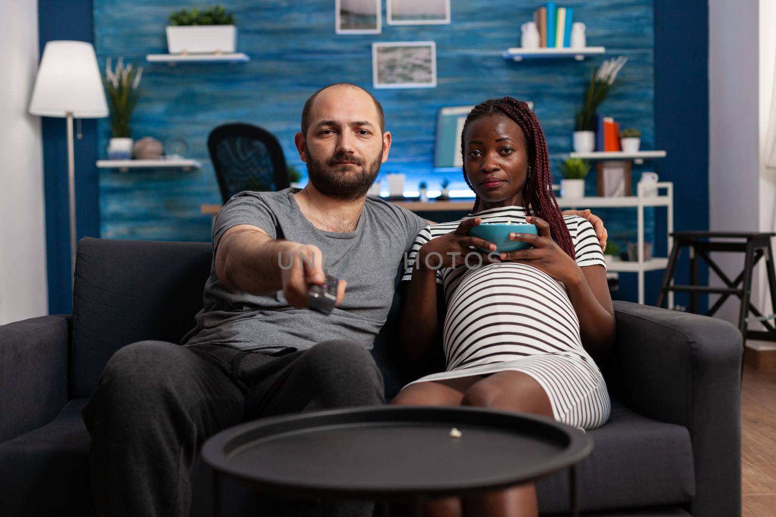 Pregnant interracial couple looking at camera watching television on sofa. POV of mixed race partners with pregnancy relaxing while eating popcorn and using TV remote control.