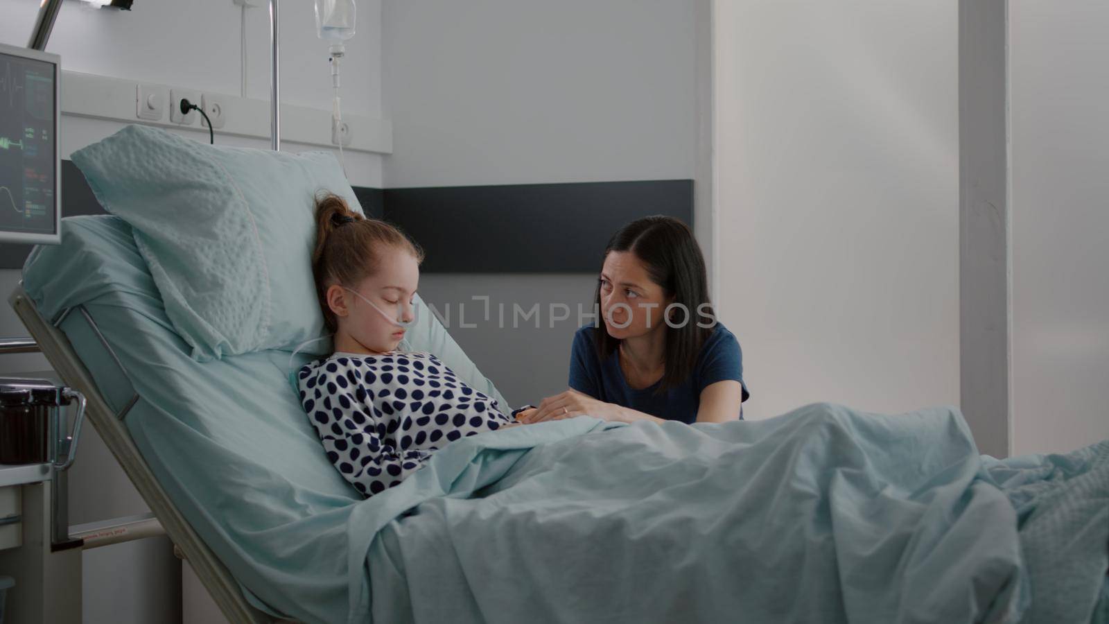 Worried mother standing with daughter praying while sleeping after recovery surgery waiting for disease expertise in hospital ward. Sick child wearing oxygen nasal tube during medicine treatment