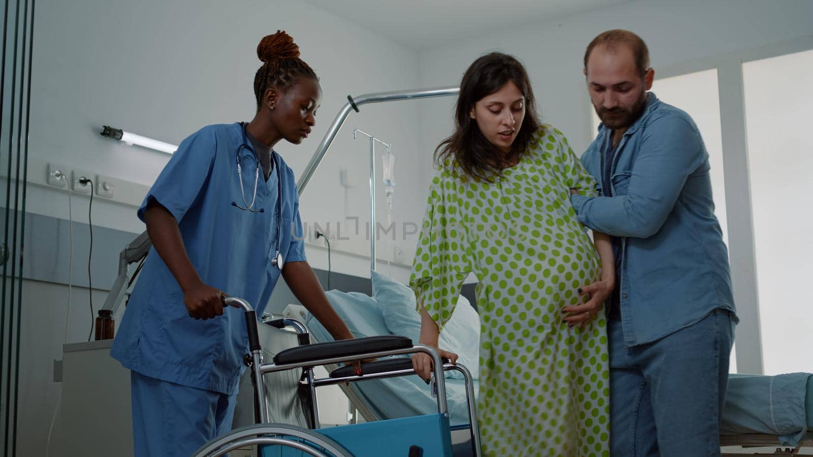 Caucasian man helping pregnant woman in hospital ward and african american nurse bringing wheelchair for patient. Young person expecting child at maternity clinic preparing for delivery