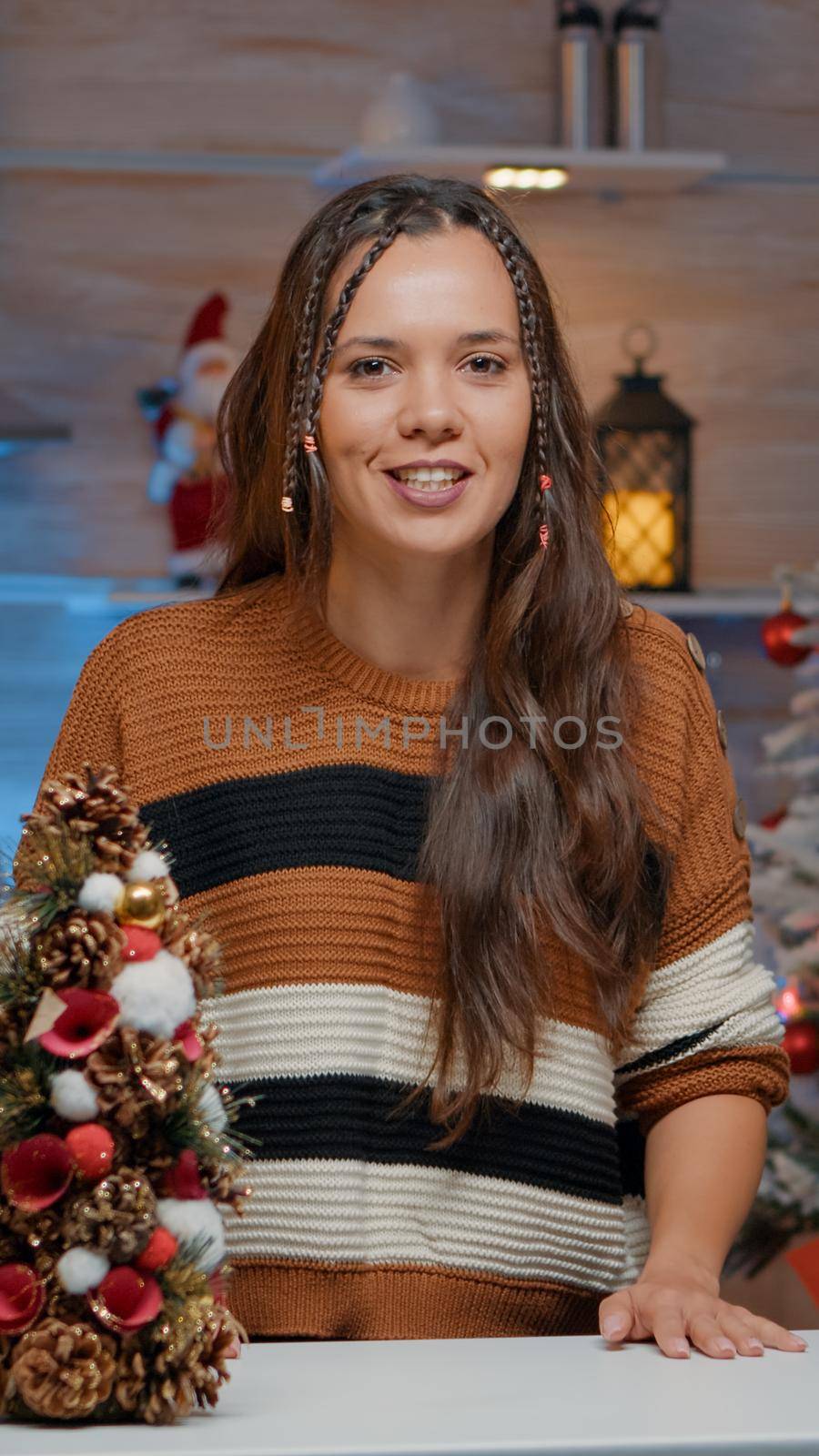 Young adult waving at video call camera in festive kitchen decorated for christmas season. Woman with sweater preparing tea while sitting in room with winter ornaments and decorations