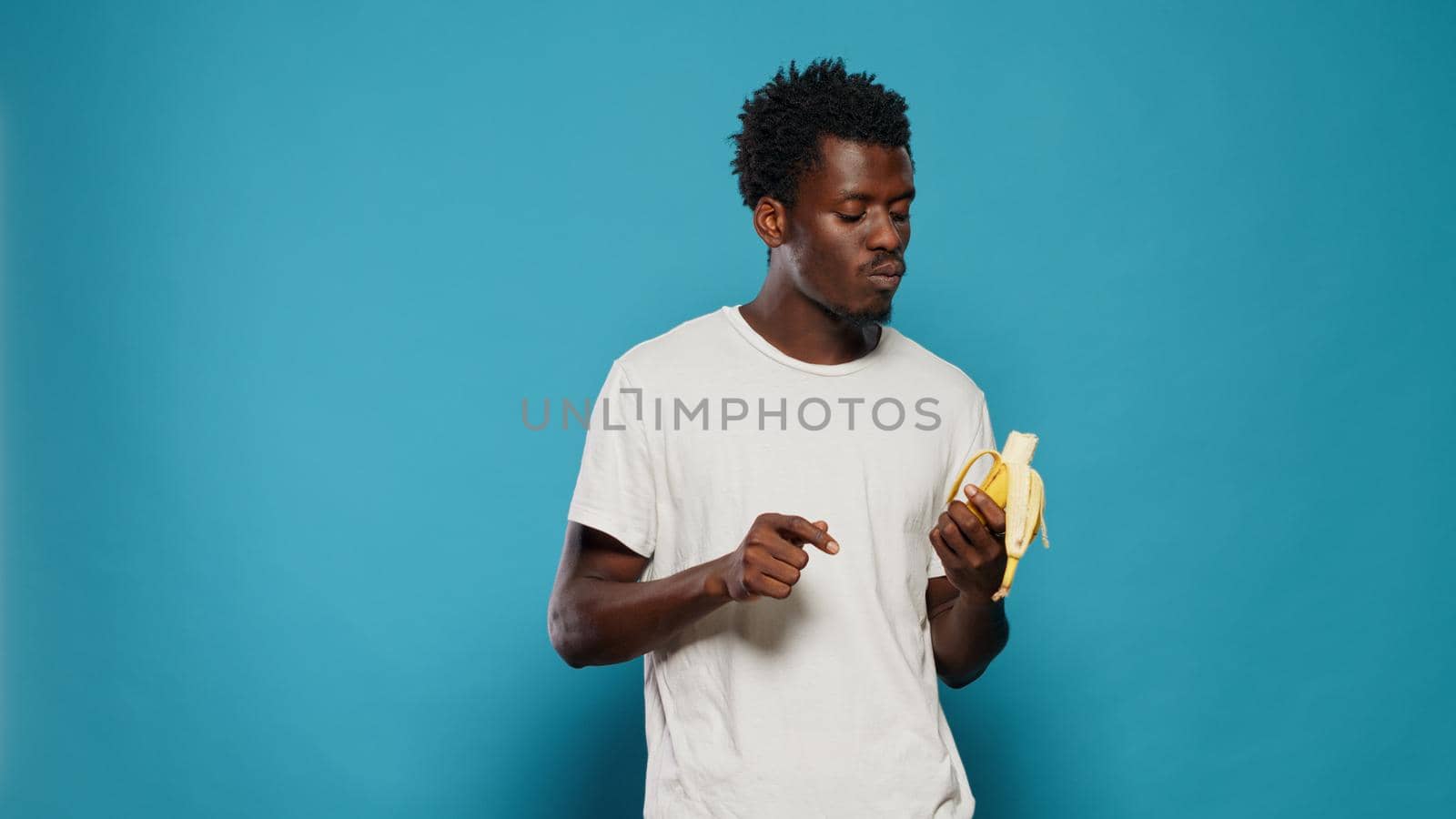 Portrait of casual adult eating banana on camera in studio. Young person enjoying yellow fruit with vitamins for healthy nutrition and fresh diet. Vegetarian man tasting natural snack