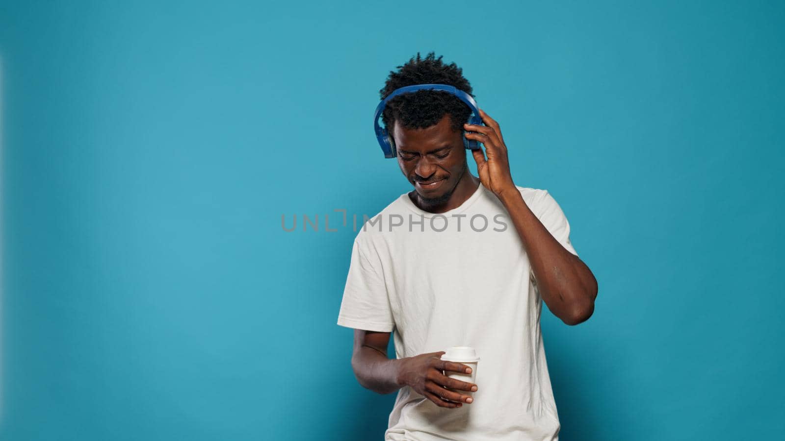 Portrait of joyful adult listening to music on headphones in studio. Casual man enjoying song rhythm while using headset and standing over isolated background. Person dancing on sound