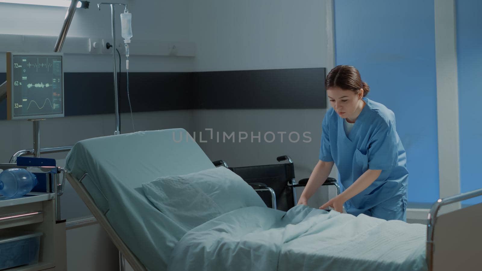 Caucasian nurse making hospital ward bed for use by sick person at modern medical facility unit. Emergency room with equipment, oxygen tube and wheelchair for intensive care treatment